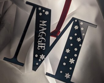 Peut inclure: Ornement de Noël en forme de lettre M en acrylique noir avec un ruban rouge et le nom Maggie inscrit dessus. La lettre est décorée de flocons de neige blancs.