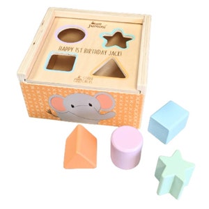May include: A wooden shape sorter toy with a light wood finish. The box has cutouts in the shape of a circle, square, star, and triangle. The box is decorated with an elephant and the text "HAPPY 1ST BIRTHDAY JACKI". Includes five colourful shapes.
