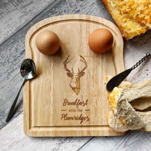 May include: A wooden breakfast serving board shaped like a slice of bread with two indentations for eggs. The board has an engraved stag head and the text "Breakfast with the Plumridges".