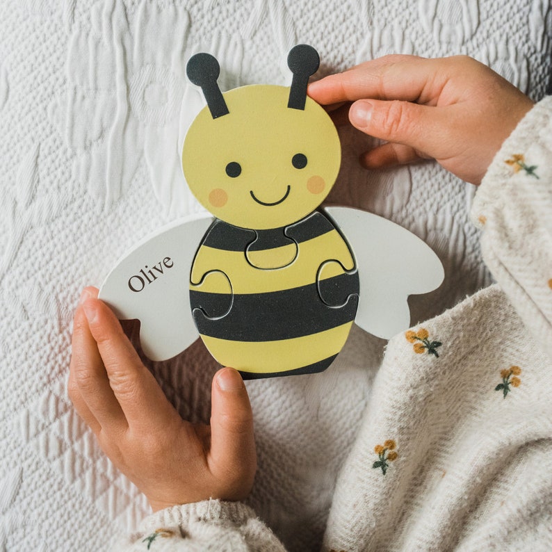 May include: A wooden puzzle of a bee with black and yellow stripes. The puzzle is in the shape of a bee with wings and a smiling face. The puzzle is personalized with the name "Olive".