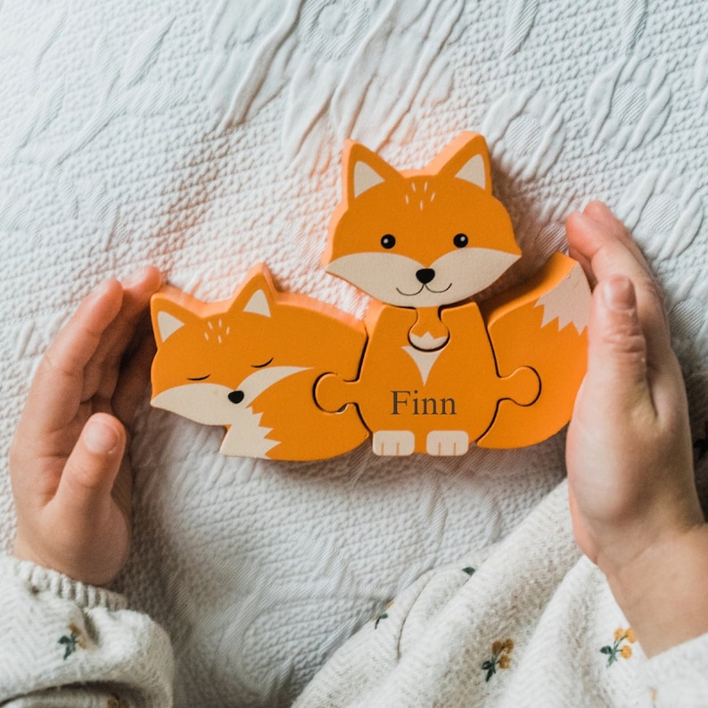 May include: A wooden fox puzzle with four pieces. The puzzle is orange and has a cute fox face on the front piece. The name "Finn" is written on the bottom piece of the puzzle.