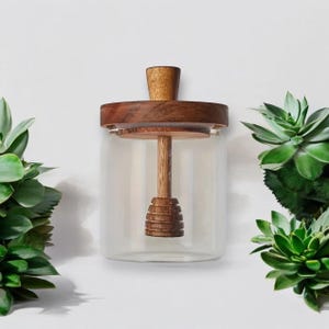 May include: A clear glass honey jar with a wooden lid and a wooden honey dipper. The jar is sitting on a white surface.