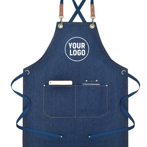May include: A blue denim apron with a pocket and the text "YOUR LOGO" in a white circle. The apron has brown leather straps and blue ties. Suitable for kitchen or workshop use.