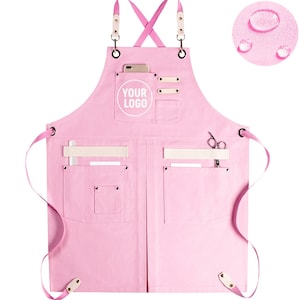 Può includere: Grembiule in tela rosa con bretelle regolabili, più tasche e un cerchio bianco con il testo "YOUR LOGO".