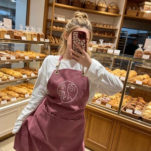 May include: A bakery scene with a variety of baked goods displayed in glass cases and on wooden shelves. A person wearing a maroon apron with a white graphic is in the foreground. Baskets and loaves of bread are visible.