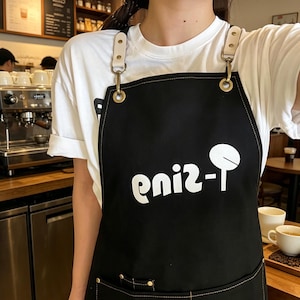 May include: Black apron with white text and a logo. The apron has adjustable straps and is worn over a white t-shirt. The background shows a coffee shop with coffee-making equipment and tables.