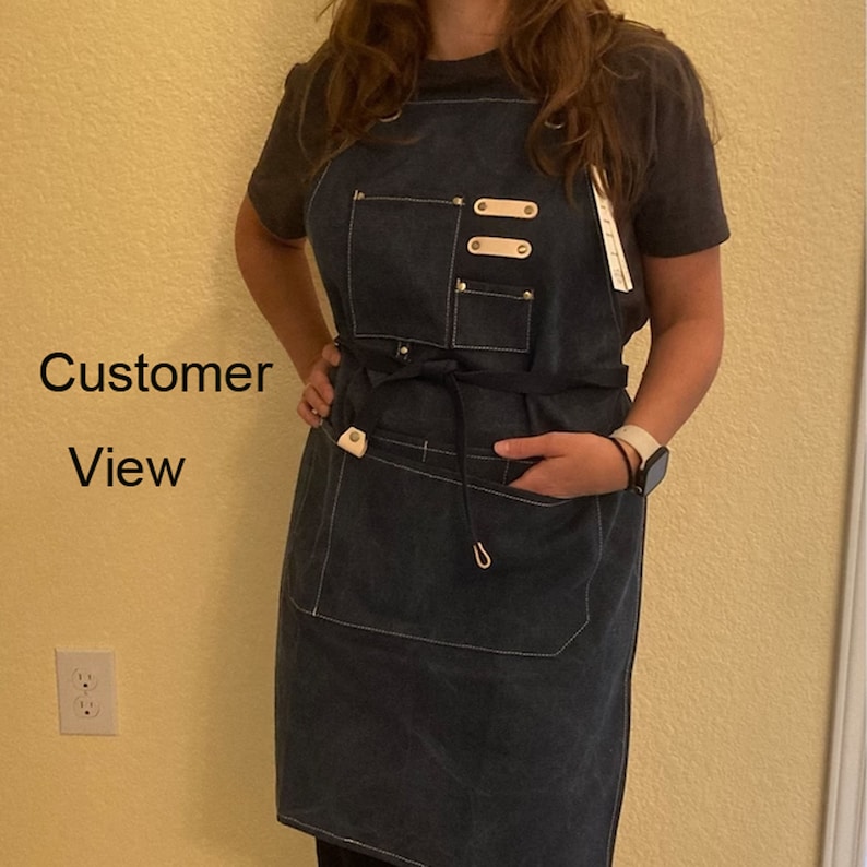May include: A dark blue denim apron with a black adjustable tie, two pockets, and three leather straps with metal rivets. The apron has white stitching and a white leather loop on the bottom.