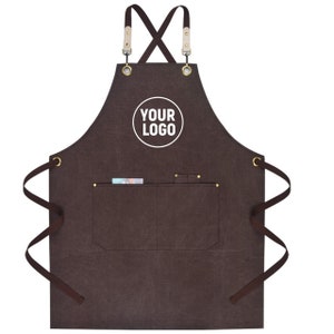 May include: Brown apron with a pocket and the text "YOUR LOGO" in a white circle. The apron has brown straps and leather accents at the top. The apron has two pockets and brass-colored rivets.