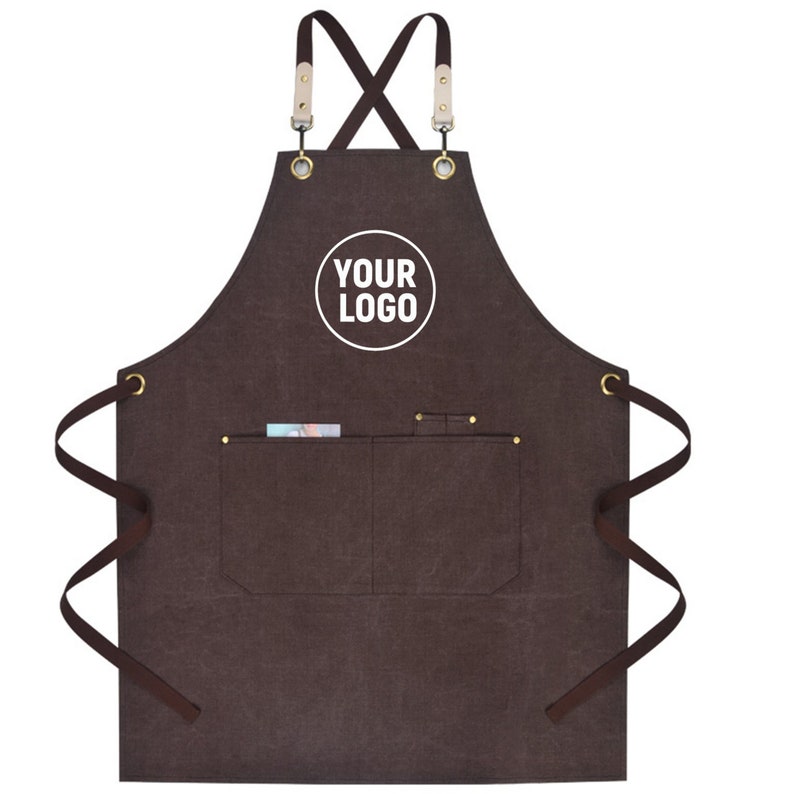May include: Brown apron with a pocket and the text "YOUR LOGO" in a white circle. The apron has brown straps and leather accents at the top. The apron has two pockets and brass-colored rivets.