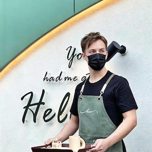 May include: A person wearing a black face mask and apron holds a tray with a mug and pastry. The apron is olive green with white stitching and a pocket. The wall behind has the words "You had me at Hello" in black script.