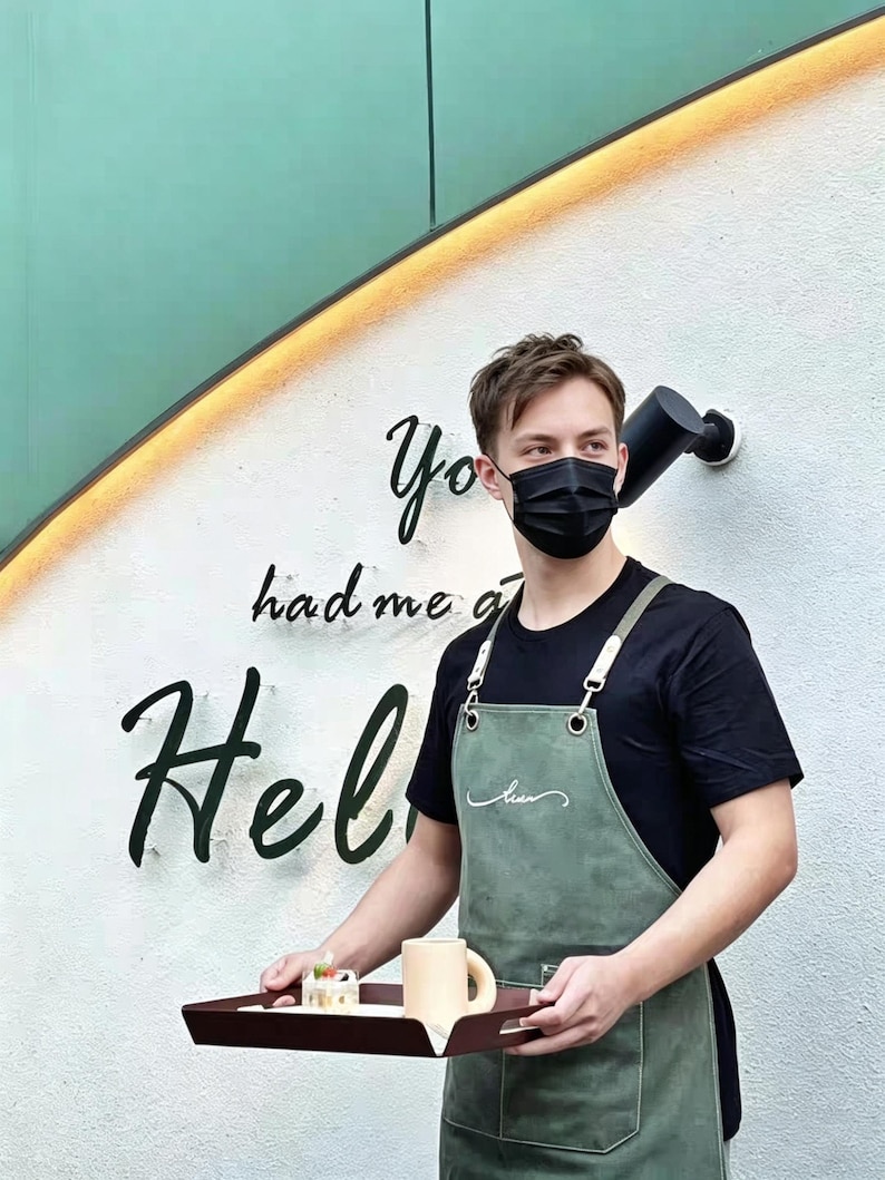 May include: A person wearing a black face mask and apron holds a tray with a mug and pastry. The apron is olive green with white stitching and a pocket. The wall behind has the words "You had me at Hello" in black script.