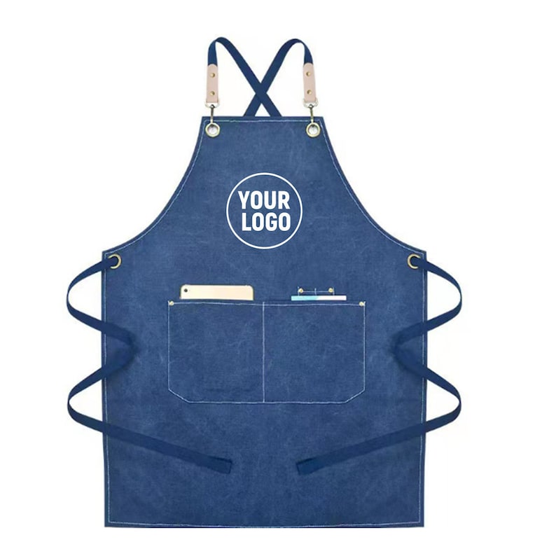 May include: A blue denim apron with a pocket and the text "YOUR LOGO" in a white circle. The apron has a cross-back design with leather straps and brass grommets. A phone and pens are in the pocket.