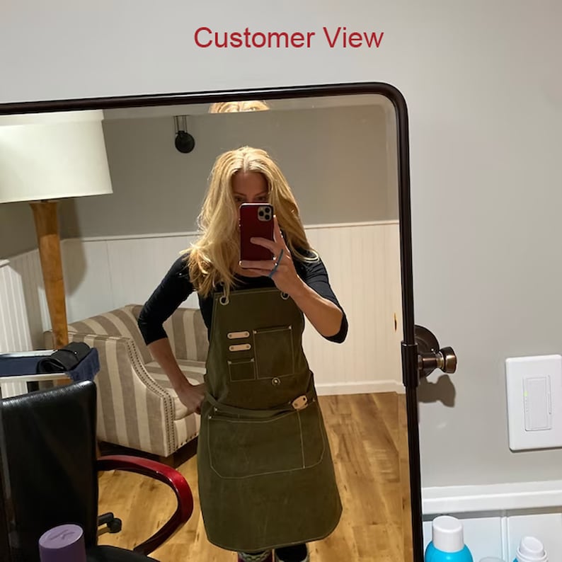 May include: A woman wearing a green canvas apron with multiple pockets and leather accents. The apron has a brown leather belt with a metal buckle. The woman is holding a red smartphone in front of her face.