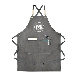 May include: A charcoal gray apron with a large front pocket and a smaller pocket. The apron has a cross-back design with leather and metal accents. The text "YOUR LOGO" is printed on the apron. Scissors, pens, and a small notepad are in the pockets.