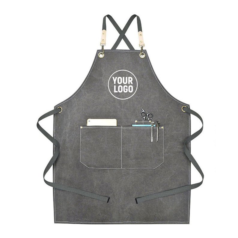 May include: A charcoal gray apron with a large front pocket and a smaller pocket. The apron has a cross-back design with leather and metal accents. The text "YOUR LOGO" is printed on the apron. Scissors, pens, and a small notepad are in the pockets.