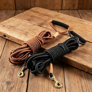 Puede incluir: Dos correas para perros, una marrón y otra negra, enrolladas sobre una superficie de madera. Cada correa tiene un asa con un agarre de espuma negra y un cierre metálico color latón. La correa marrón es de un tono más claro que la negra.