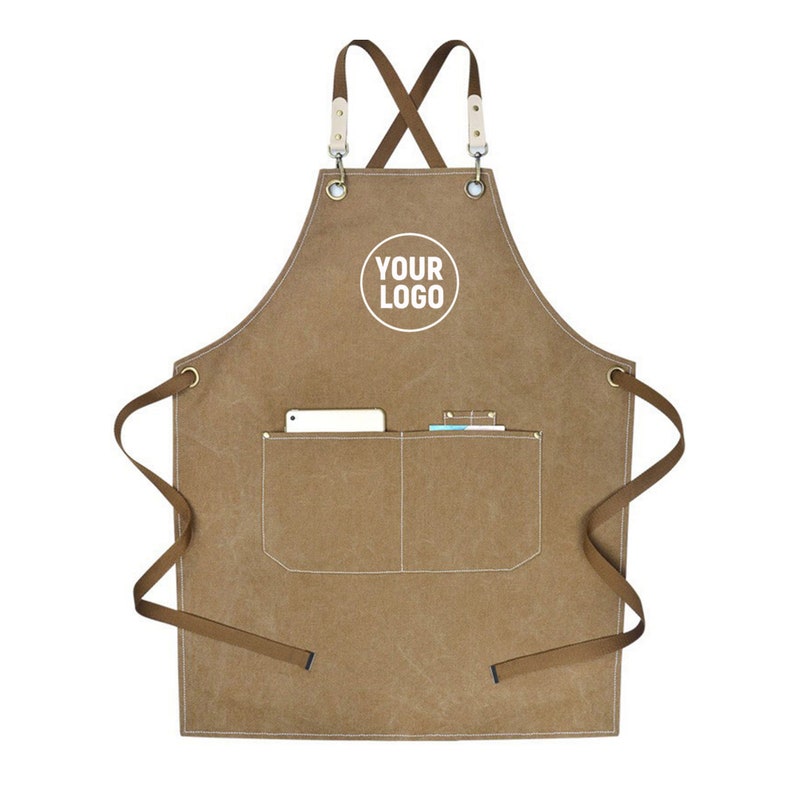 May include: A brown canvas apron with a white pocket and the text "YOUR LOGO" in a white circle. The apron has brown straps and a pocket containing a phone and a small notebook. The apron is designed for use in a kitchen or workshop.