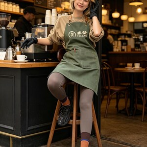 May include: A green apron with the text "TAIZAN" and a logo, worn by a person in a coffee shop. The apron has brown leather straps. The person is wearing black gloves and is sitting on a wooden stool.
