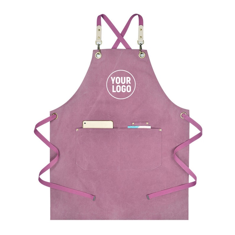 May include: A dusty rose apron with a pocket, cross-back straps, and a white circle with the text "YOUR LOGO". The apron has a pocket containing a phone and small notepad. The straps are a lighter pink color.