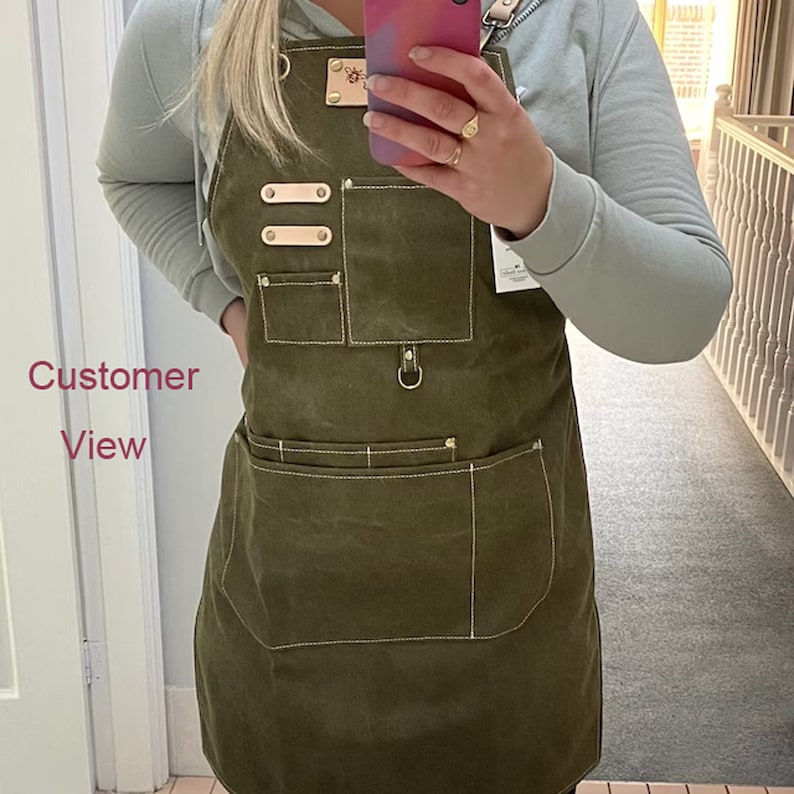 May include: A green canvas apron with multiple pockets and a leather strap. The apron has a metal ring on the front and three brass rivets on the left side.