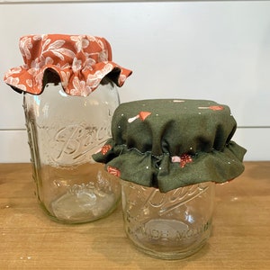 May include: Two glass jars with fabric lids. The larger jar has a lid with a floral pattern in shades of orange and white. The smaller jar has a lid with a mushroom pattern in shades of green and pink.