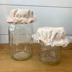 May include: Two clear glass mason jars with fabric daisy-covered lids. The lids are a light peach color with white daisies and a white lace trim. The jars are empty and sit on a wooden surface.