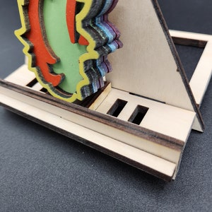 Può includere: Un supporto per telefono in legno con un design multistrato e colorato. Il supporto ha una fessura per un telefono e un piccolo ripiano sul retro.
