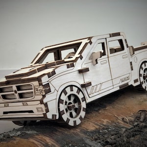 Op de afbeelding: Een wit houten model van een pick-up truck met zwarte banden en een zwarte streep aan de zijkant. De truck heeft een zwarte grille en koplampen. Het model staat op een stuk hout.