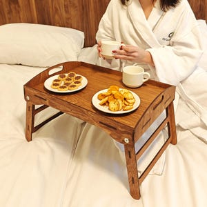 Puede incluir: Una bandeja de desayuno de madera con patas plegables, que contiene dos platos de comida y dos tazas blancas. La bandeja es de color marrón cálido y está colocada sobre una cama blanca. La bandeja tiene asas.