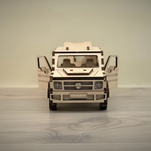 K&ouml;nnte beinhalten: Ein Holzmodell eines Mercedes-Benz G-Wagens mit ge&ouml;ffneten T&uuml;ren. Das Modell ist aus hellem braunem Holz gefertigt und verf&uuml;gt &uuml;ber einen detaillierten Innenraum.