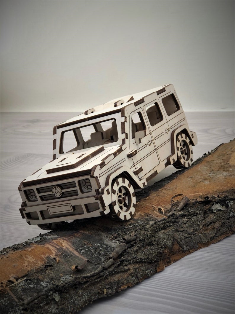 K&ouml;nnte beinhalten: Ein wei&szlig;es Holzmodell eines Mercedes-Benz G-Klasse SUV mit braunen Akzenten, das auf einem St&uuml;ck Baumrinde platziert ist.