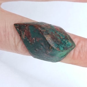 Pode incluir: Um anel de declaração verde e marrom feito de um material único e texturizado. O anel tem a forma de um oval pontiagudo e tem uma aparência áspera e orgânica.