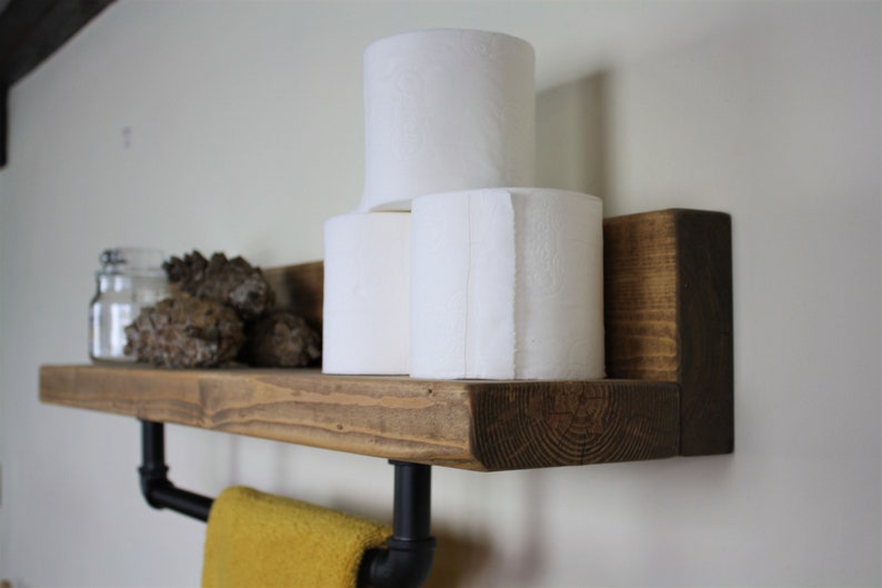 Pu&ograve; includere: Uno scaffale in legno rustico con un portasciugamani in tubo nero e tre rotoli di carta igienica. Lo scaffale &egrave; montato su una parete bianca.