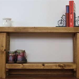 May include: A rustic wooden bench with a shelf, featuring a white ceramic candle warmer on the top shelf and a pair of colourful boots on the lower shelf.  The bench is decorated with a set of bookends shaped like bicycles holding up two books.