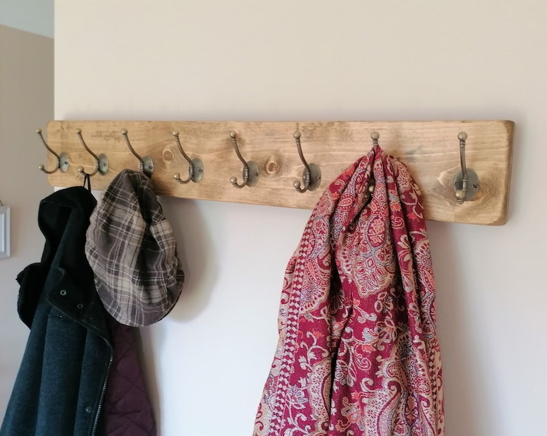 May include: A wooden coat rack with six hooks, featuring a rustic brown finish. A red and cream patterned scarf hangs from one hook, a plaid hat from another, and a black jacket from a third.