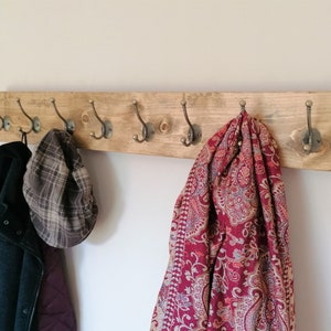 May include: A wooden coat rack with six hooks, featuring a rustic brown finish. A red and cream patterned scarf hangs from one hook, a plaid hat from another, and a black jacket from a third.