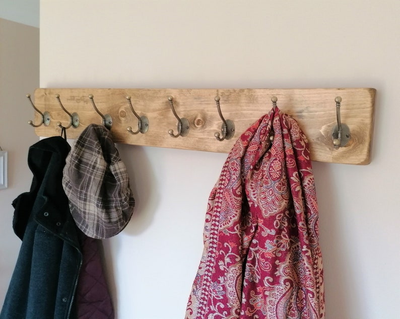 May include: A wooden coat rack with six hooks, featuring a rustic brown finish. A red and cream patterned scarf hangs from one hook, a plaid hat from another, and a black jacket from a third.