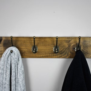 Puede incluir: Un perchero de madera r&uacute;stico con cinco ganchos de lat&oacute;n antiguo. El perchero est&aacute; te&ntilde;ido de un color marr&oacute;n oscuro y tiene un acabado desgastado. Una bufanda de punto blanca y una chaqueta negra cuelgan de los ganchos.