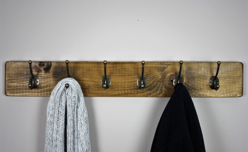 Puede incluir: Un perchero de madera r&uacute;stico con cinco ganchos de lat&oacute;n antiguo. El perchero est&aacute; te&ntilde;ido de un color marr&oacute;n oscuro y tiene un acabado desgastado. Una bufanda de punto blanca y una chaqueta negra cuelgan de los ganchos.