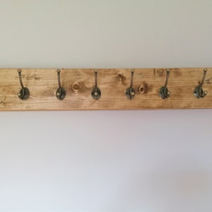 May include: A wooden coat rack with eight antique brass hooks. The rack is stained a light brown color and has a rustic look.