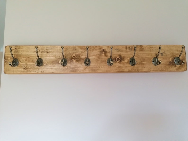 May include: A wooden coat rack with eight antique brass hooks. The rack is stained a light brown color and has a rustic look.