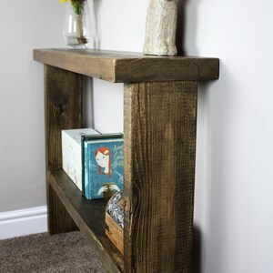 May include: A rustic wooden shelf unit with two shelves. The shelf is stained a dark brown color and has a distressed finish. The top shelf has a white ceramic rabbit figurine on it. The bottom shelf has two books and a small wooden box on it.