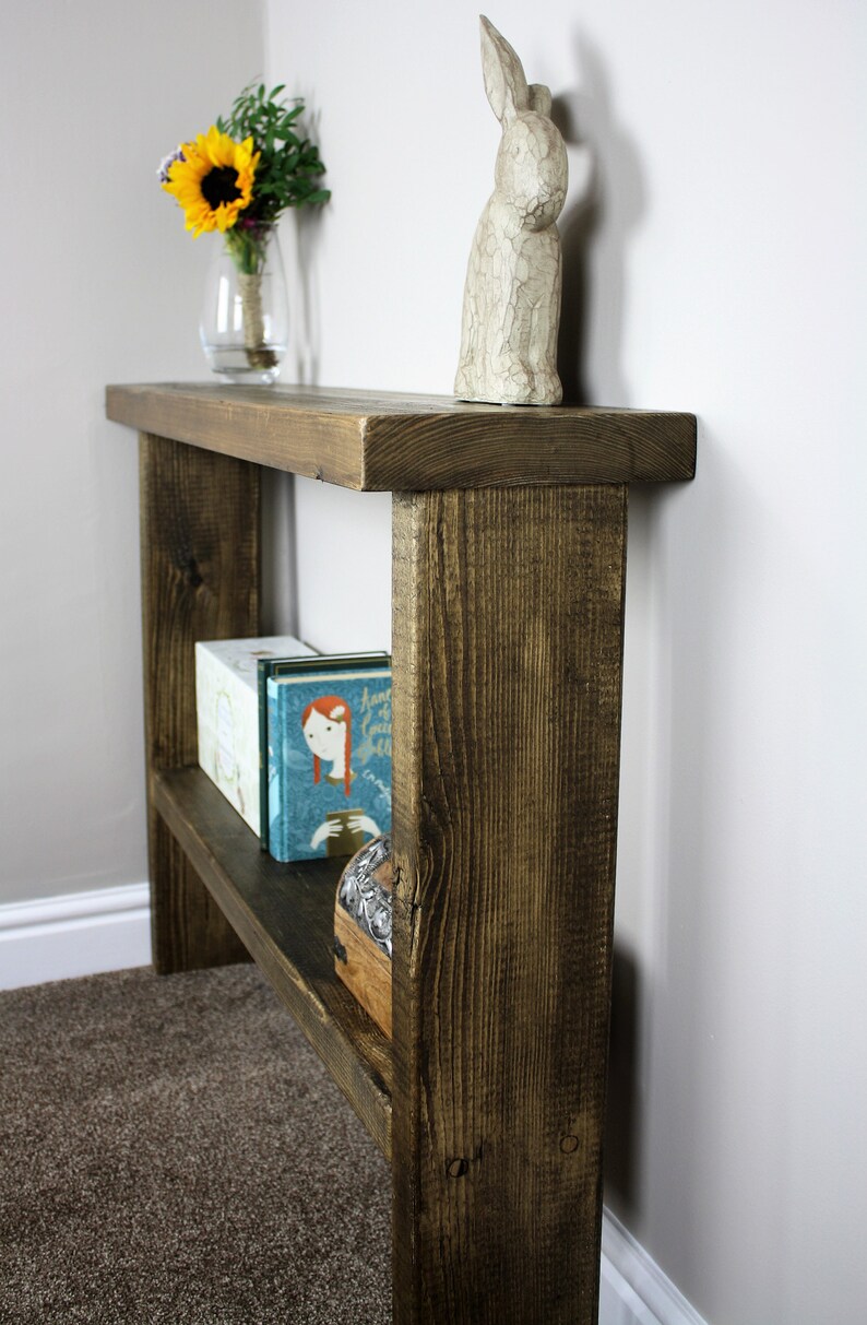 May include: A rustic wooden shelf unit with two shelves. The shelf is stained a dark brown color and has a distressed finish. The top shelf has a white ceramic rabbit figurine on it. The bottom shelf has two books and a small wooden box on it.