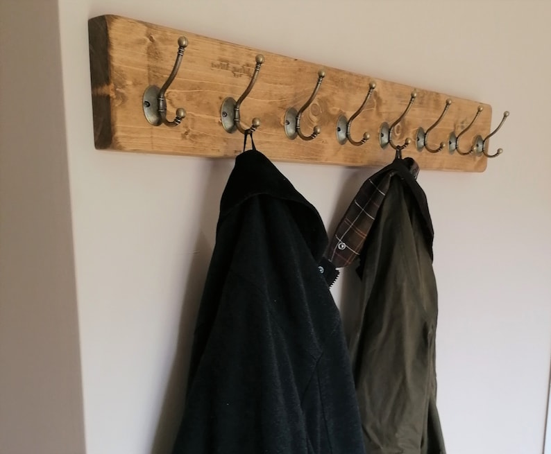 May include: A rustic wooden coat rack with multiple hooks, featuring a natural wood finish and antique brass hooks. Two coats are hanging on the rack, one black and one green.