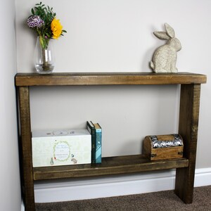 May include: A rustic wooden console table with a shelf. The table has a dark brown stain and is made of reclaimed wood. It features a decorative ceramic rabbit figurine on the top shelf and a small wooden box on the lower shelf.
