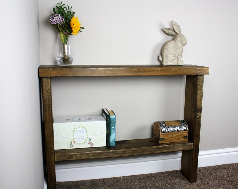 May include: A rustic wooden console table with a shelf. The table has a dark brown stain and is made of reclaimed wood. It features a decorative ceramic rabbit figurine on the top shelf and a small wooden box on the lower shelf.