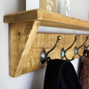 Peut inclure: &Eacute;tag&egrave;re en bois rustique avec un support triangulaire et cinq crochets en m&eacute;tal. L'&eacute;tag&egrave;re est teint&eacute;e d'une couleur brun clair et a une finition vieillie. Une veste noire et une &eacute;charpe &agrave; motifs rouge et blanc sont accroch&eacute;es aux crochets.