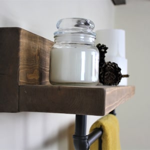 Pu&ograve; includere: Uno scaffale in legno rustico con un portasciugamani in tubo metallico nero sotto. Lo scaffale ha una candela bianca in un barattolo di vetro e una pigna in cima. Un asciugamano giallo &egrave; appeso al portasciugamani.
