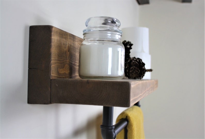 Pu&ograve; includere: Uno scaffale in legno rustico con un portasciugamani in tubo metallico nero sotto. Lo scaffale ha una candela bianca in un barattolo di vetro e una pigna in cima. Un asciugamano giallo &egrave; appeso al portasciugamani.