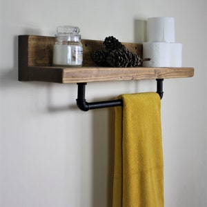Pu&ograve; includere: Uno scaffale in legno rustico con un portasciugamani in tubo di metallo nero. Lo scaffale &egrave; decorato con un barattolo di vetro, pigne e due rotoli di carta igienica.